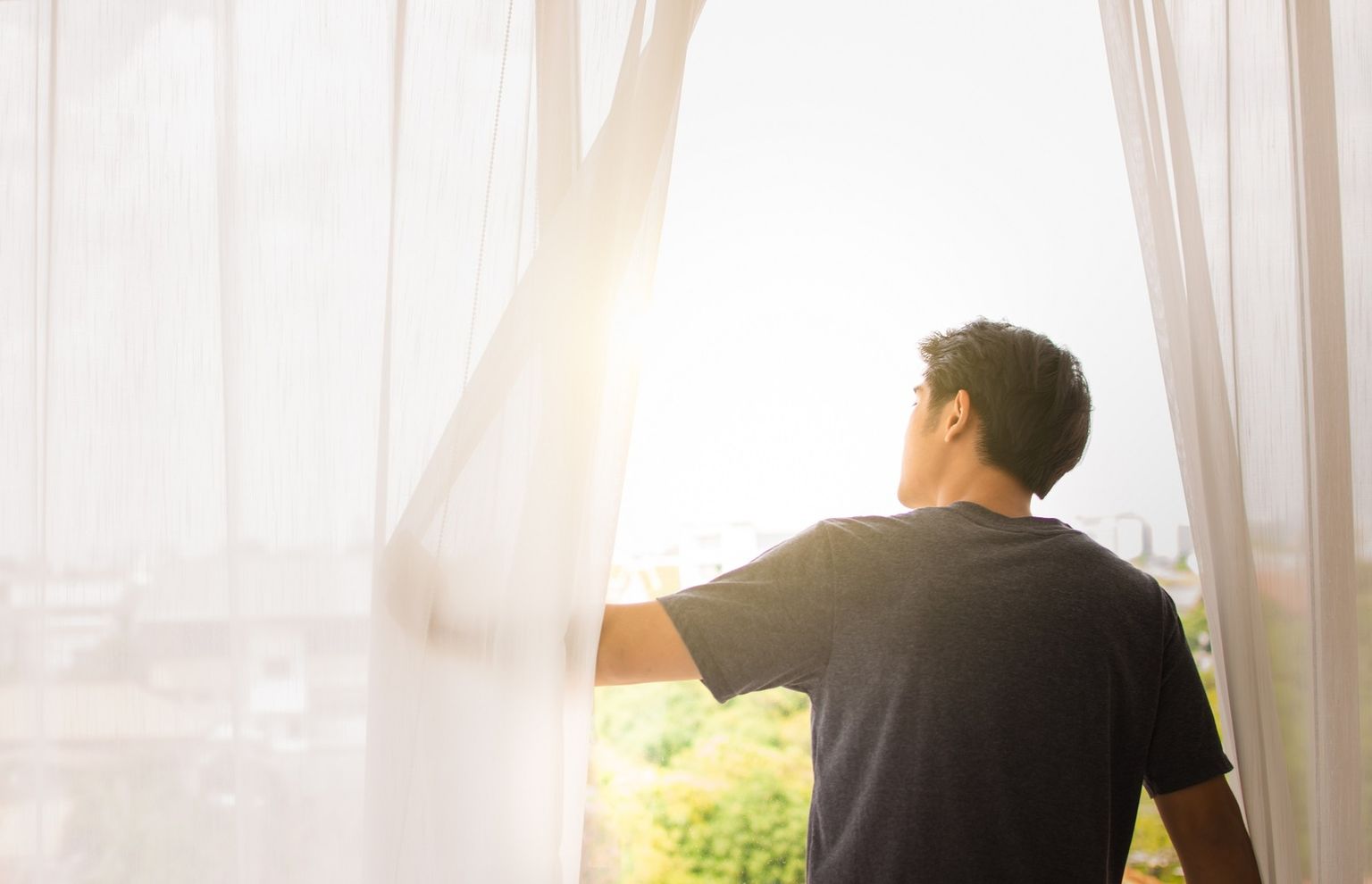 One person pushes aside a light window curtain to allow the sun to shine through.